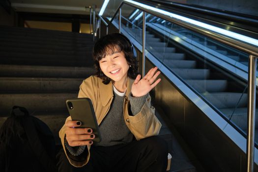 Portrait of korean girl sits on stair and waves hand at mobile phone, video chats on smartphone, greets friend on social media.
