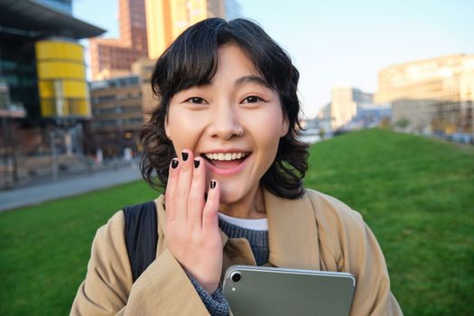 Image of korean girl with happy face, walks around town with student tablet, stands on street and smiles. Copy space