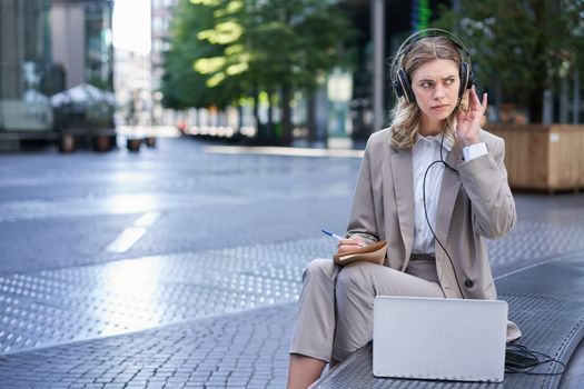 Woman sitting on a street with laptop and headphones plugged in, taking notes. Corporate worker attend online team meeting and writing down information, working outdoors.