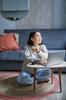 Vertical shot of asian girl sits on floor at home, working on laptop, studying at cozy place, using computer to freelance.