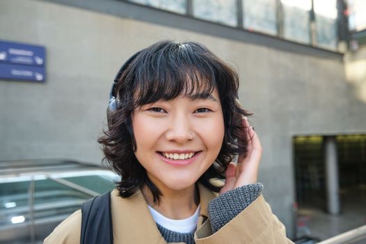 Close up of smiling brunette girl in headphones, listens music, travels around city, commutes to work, stands on a street.