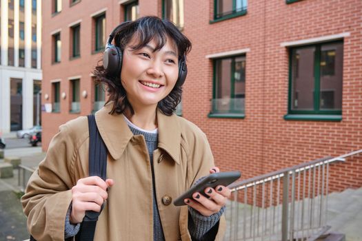 Happy young woman stands on street with backpack and smartphone, listens music in headphones, waits someone outdoors, travels around city.