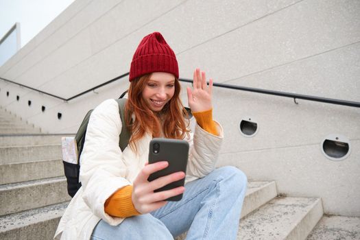 Happy college girl talks with friends on video chat smartphone app, sits on stairs outdoors uses her mobile phone application, waves hand at telephone camera.