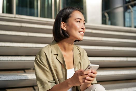 People and technology. Smiling beautiful asian woman sitting on stairs in city, holding mobile phone. Girl with smartphone rests outside.