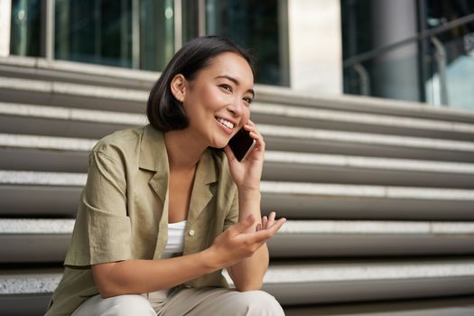 Portrait of beautiful asian girl talks on mobile phone, sits on street stairs. Woman with smartphone smiling, making a call.