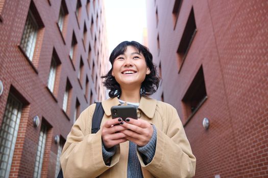 Lower angle view of brunette korean girl, listening music in headphones, walking along street and looking at smartphone, reading message on mobile phone.