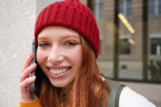 Young people and mobile connection. Happy redhead girl talks on phone, makes telephone call, stands outdoors with backpack and uses smartphone app.