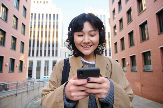 Portrait of cute girl tourist, korean woman in headphones, looks at mobile phone, uses smartphone app, map or text messages, listens music in headphones.