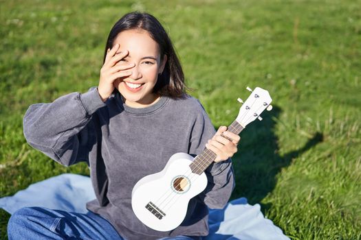 Smiling asian girl with ukulele, playing in park and singing, lifestyle concept. Copy space