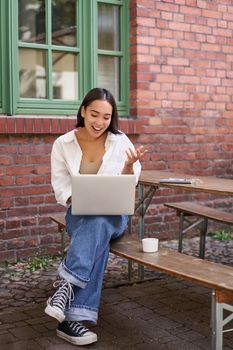 Vertical shot of asian girl with laptop, talking on video call, chatting online, sitting in cafe outdoors. People and communication concept
