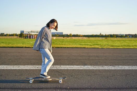 Beautiful asian skater girl riding her longboard on sunny empty road. Young woman enjoying her skate ride smiling and laughing.