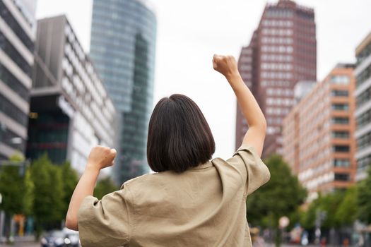 Excitement. Happy girl rear view, raising her hands up and looking at big city skyscrappers, feeling confident, cheering and triumphing.