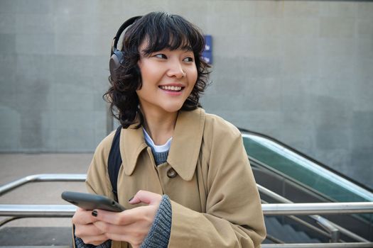 Technology and people. Young asian girl, student standing on street in headphones, listens music and smiling, chats on mobile phone, sends a text message, waits for someone on street.