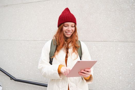 Young redhead woman with red hat, uses her digital tablet outdoors, stands on street with gadget, connects to wifi internet and searches for a location in internet.