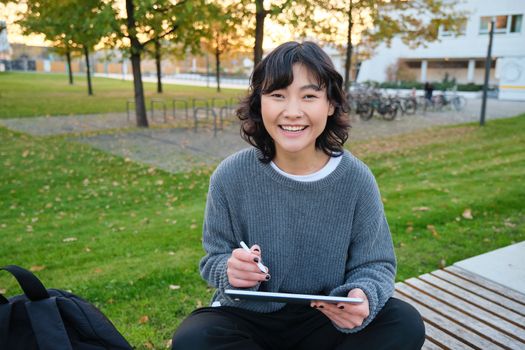 Young girl, korean artist or art student sits in park with digital tablet, draws with graphic pen, scatches a design or project, looks around.