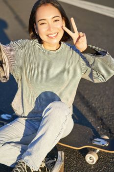 Selfie of asian girl sitting on skateboard, taking photo on smartphone, smiling and showing peace v-sign.