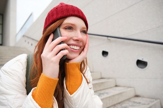 Beautiful smiling redhead female model, sits on street and talks on mobile phone, uses smartphone app to call abroad, laughing during telephone conversation.