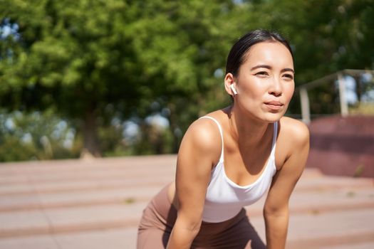 Portrait of asian woman taking break, breathing heavily and panting after running, jogger standing and wiping sweat off forehead, smiling pleased.