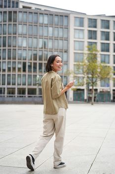 Silhouette of asian woman walking on street in wireless headphones, holding smartphone.