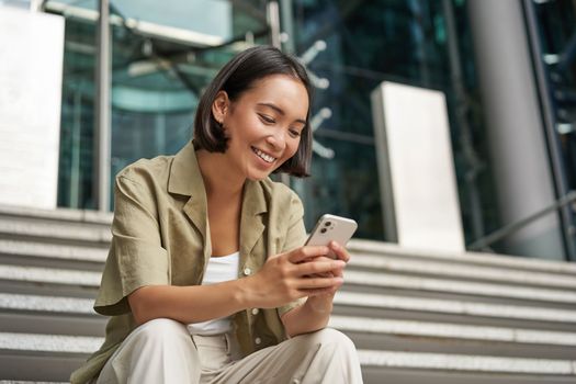 Technology and communication. Young smiling girl, asian woman sits with smartphone, reads message with big smile.