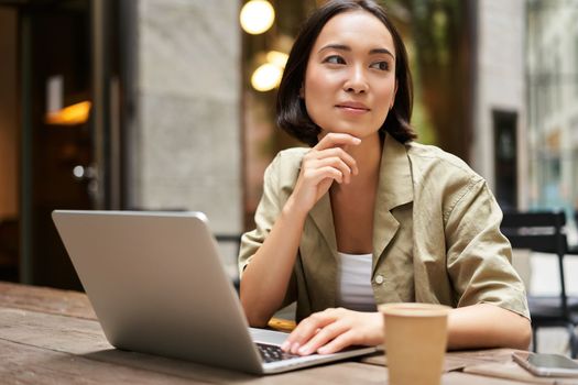 Young woman working in a cafe, using laptop and drinking coffee. Asian girl student with computer studying remotely, sitting on bench near shop.