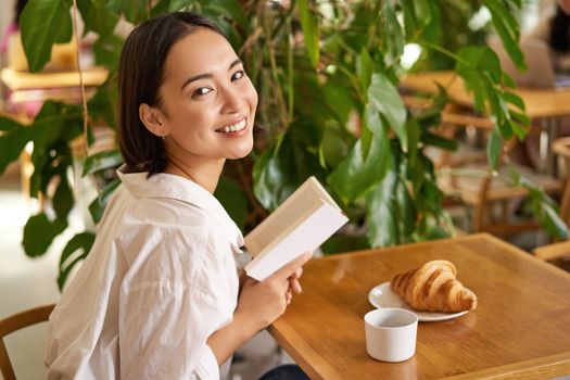 Beautiful young asian woman with a book in hands, sitting in cafe, drinking coffee and eating croissant, smiling, looking mysterious.