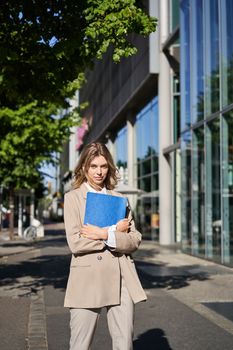 Vertical shot of businesswoman with folder, standing on a street near office buildings, wearing suit for office work.