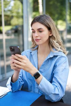 Vertical shot of successful businesswoman sitting outdoors in park, working on her project, using mobile phone. Corporate people concept.