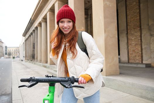 Smiling redhead european girl drives public e-scooter, tourist explores city, rides in city centre.