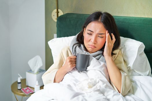 Portrait of asian woman with headache, catching col, staying on sick leave at home, lying in bed, drinking hot tea, having flu.