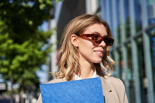 Close up portrait of smiling young woman in sunglasses, corporate suit, holding a folder with work documents, going to an interview, standing outdoors.