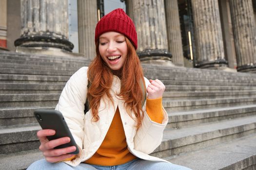 Achievement celebration. Happy redhead girl sits on stairs outdoors and looks at phone, triumphs, wins something and looks satisfied at mobile screen.