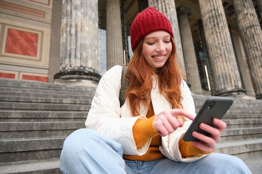 Stylish young redhead woman, talking on mobile phone app, using social media application, looking for something online on smartphone, sits on stairs outdoors.