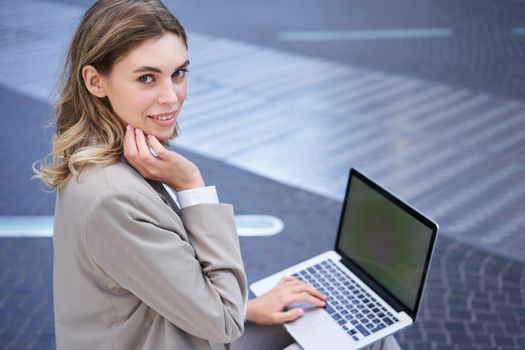 Young businesswoman using laptop while sitting outdoors in city centre, typing on keyboard. Girl preparing for interview, wearing suit.