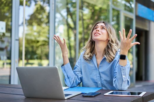 Disappointed young businesswoman feeling frustrated, sitting with laptop outdoors on street, looking up and complaining.