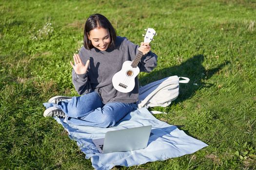 Cheerful asian girl, musician waves at her laptop, say hi to video chat, plays ukulele and records it on laptop, sits on grass in park.