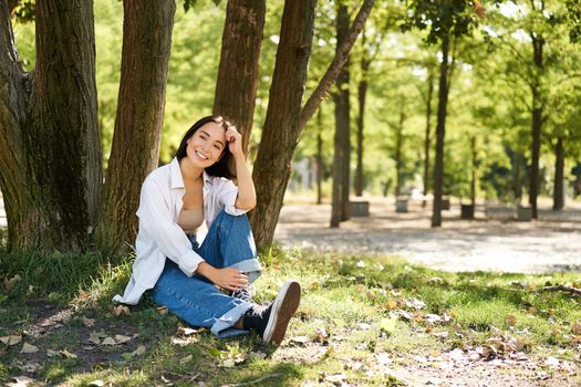 Young people. Beautiful asian girl sits near tree in park and rests, smiling and looking into distance, relaxing outdoors on fresh air in summer.