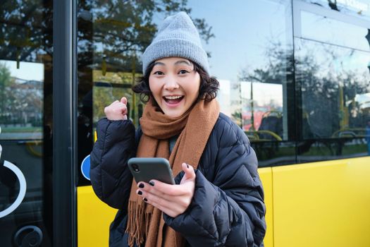 Enthusiastic girl rejoice, reads message on mobile phone and celebrates, stands near her bus on public transport stop and looks excited, posing in warm winter clothes.