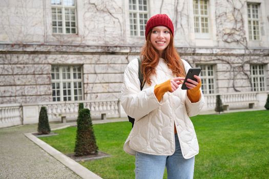 Portrait of girl tourist, wonders around city with smartphone map application, holding mobile phone and smiling, looking up info in internet.