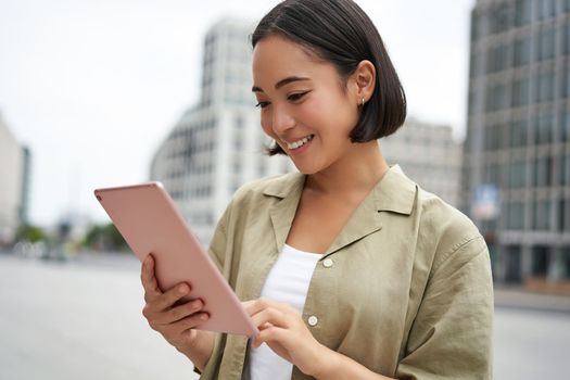 Portrait of asian woman reading, using tablet while standing on street, smiling while looking at screen.