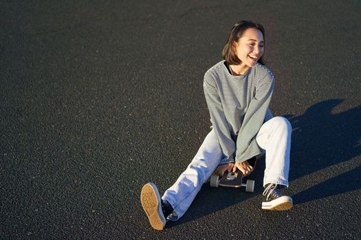 Happy beautiful korean teen girl sits on her skateboard, cruising on longboard, wearing casual clothes.