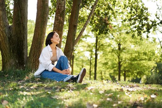 Portrait of asian girl relaxing, leaning on tree and resting in park under shade, smiling and enjoying the walk outdoors.