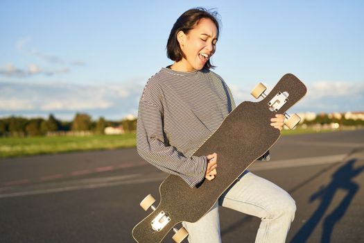 Beautiful asian teen girl playing with her longboard, holding skateboard as if playing guitar, standing on road on sunny day.