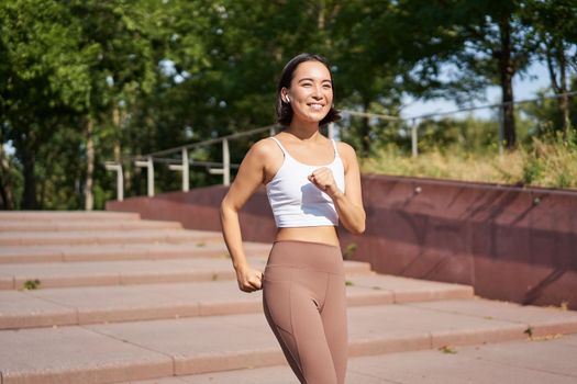 Wellbeing and sport. Asian fitness girl runner, jogging in park, running on street in leggings, smiling happily.