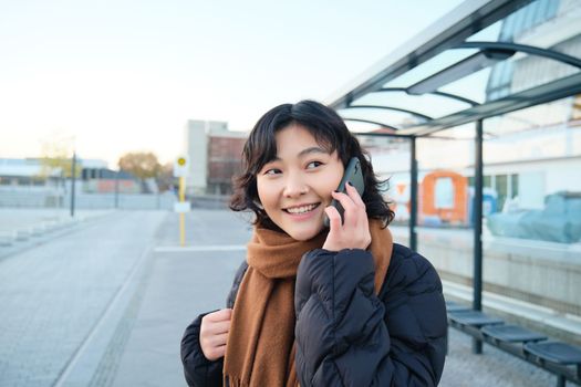 Beautiful smiling korean girl, waiting on bus stop, using public transport, talking on mobile phone, going somewhere in city.