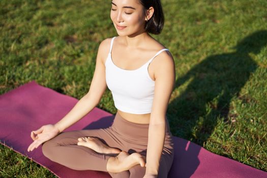 Portrait of smiling asian woman meditating, doing yoga on fresh air, relaxing on rubber mat, exercising in park, breathing air, being calm.