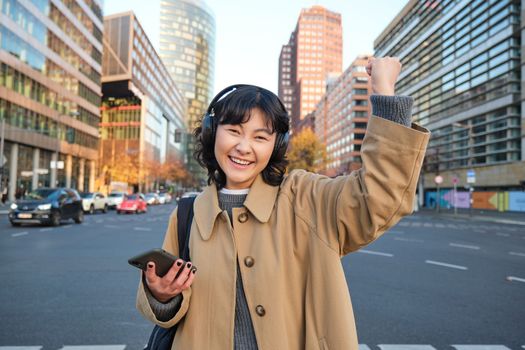 Young happy woman celebrating on street, holding smartphone and cheering, reacts amazed to good news, posing happy in city centre.