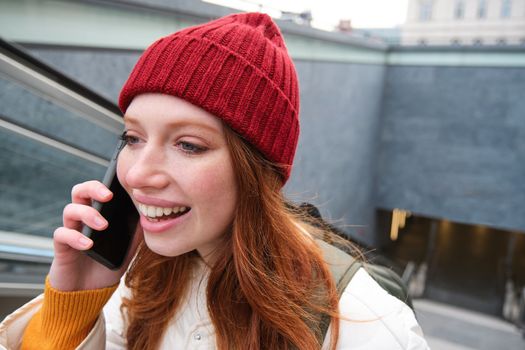 Portrait of young redhead woman walks around city, goes up stairs with mobile phone, talks on smartphone and smiles.