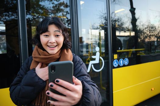 Enthusiastic asian woman, standing on bus stop with smartphone, looking at phone screen with amazed, triumphing face, winning, hear great news on video chat.