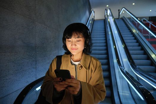 Cute young woman going down the escalator to the tube, using subway metro to commute to work or university, standing with smartphone.